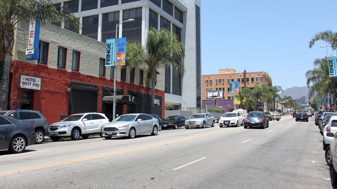 A street scene with buildings, parked cars, palm trees, and a clear sky. One building has a sign for a hotel.