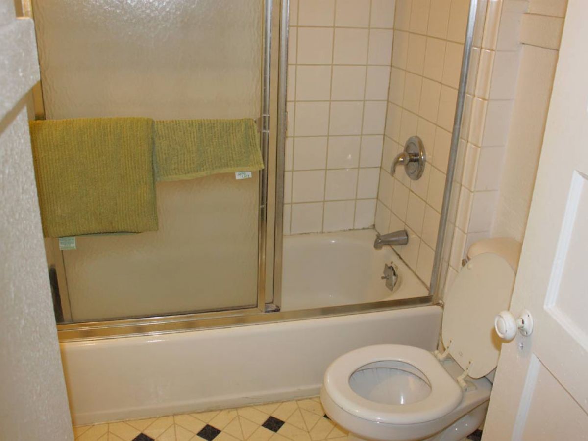 This image shows a bathroom with a bathtub, a shower door, green towel, a toilet with the lid up, and tiled walls and floor.