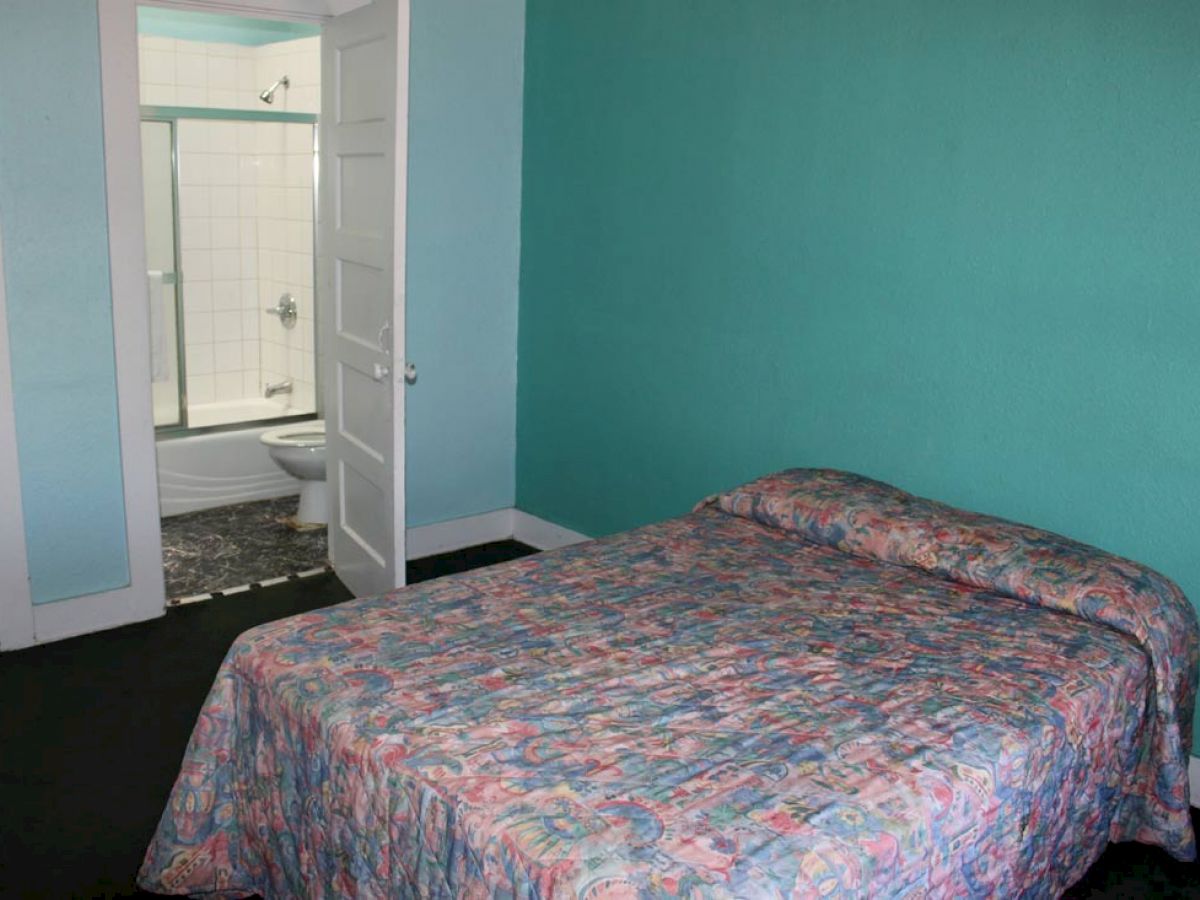 A simple bedroom with a bed covered in a patterned bedspread. The room has a turquoise wall and an open door leading to a small bathroom.