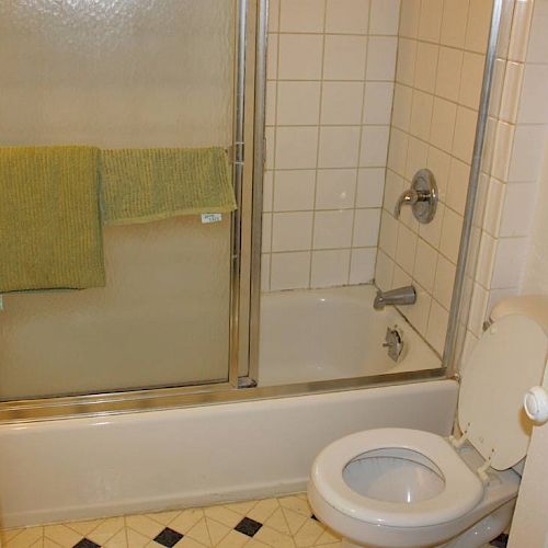 The image shows a bathroom with a shower-tub combo, a glass sliding door, a green towel, white tiled walls, a toilet with the lid up, and a door.