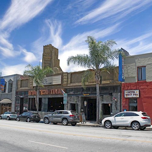 A street view featuring a row of buildings, several cars parked along the sidewalk, and palm trees under a partly cloudy sky.