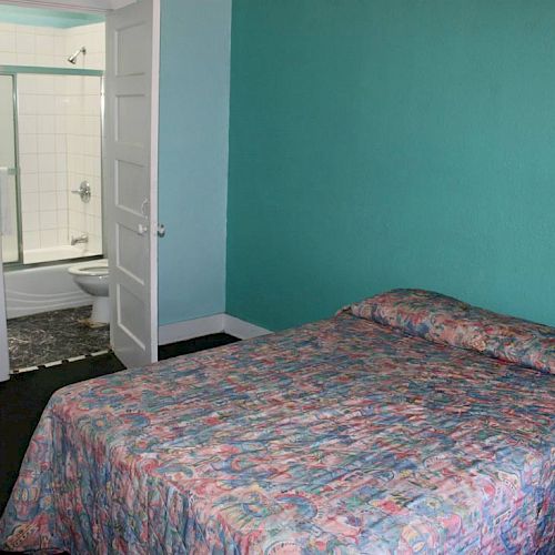 The image shows a simple bedroom with a bed against a teal wall, and an open door leading to a small, white-tiled bathroom.