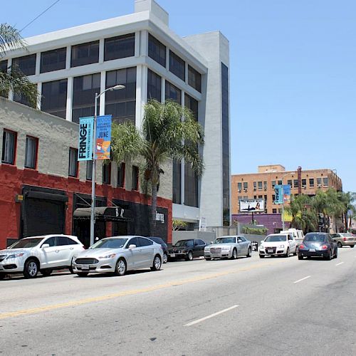 A city street with parked cars, buildings, palm trees, and a clear sky.