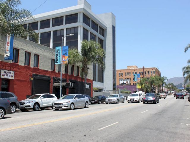 A street view of a hotel with a red and white facade, palm trees, and a busy street with cars.