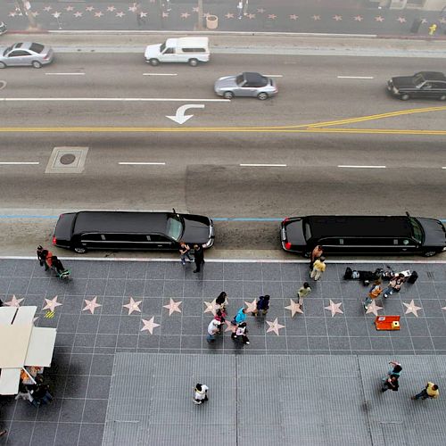 An overhead view of a street with traffic, two limousines parked, and people walking on a sidewalk with star plaques embedded, ending the sentence.