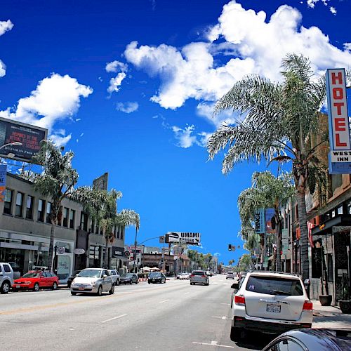 A busy street with cars, palm trees, and various shops. A large blue sky with white clouds is seen above, and a hotel sign is visible on the right.