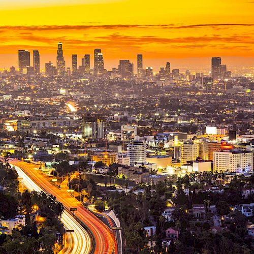 A scenic city skyline during sunset with a bustling freeway leading into the urban area; tall buildings illuminate the horizon, creating a vibrant view.