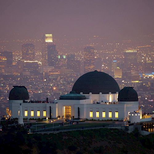 The image shows the Griffith Observatory prominently with the cityscape of Los Angeles illuminated in the background during twilight.