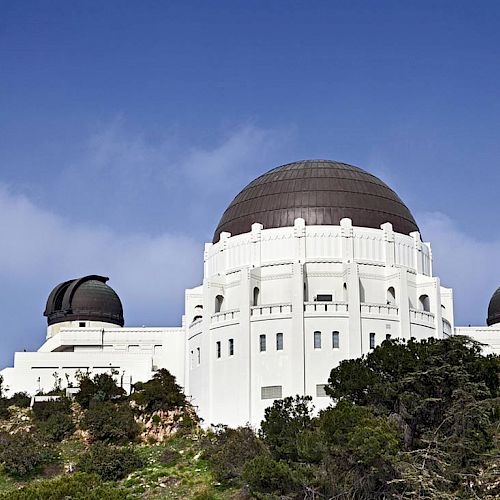 This image shows a large white astronomical observatory with a prominent dome, surrounded by greenery and set against a clear blue sky.