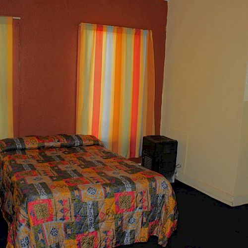 The image shows a small room with a bed, colorful striped curtains, a chair, a small table, and a wall-mounted sink.