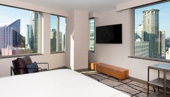 The image shows a modern bedroom with large windows offering a city view, a wall-mounted TV, a bed, a chair, and a small table.