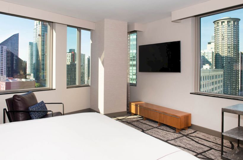 The image shows a modern bedroom with large windows offering a city view, a wall-mounted TV, a bed, a chair, and a small table.
