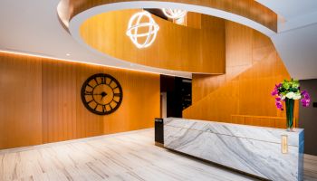 A modern lobby features a spiral wooden staircase, a large clock on the wall, a marble reception desk with flowers, and stylish light fixtures.