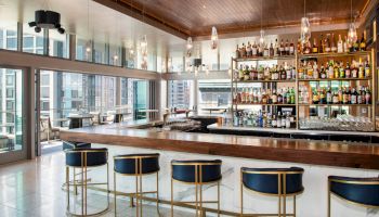 A modern bar with a wooden countertop, high-back bar stools, glass shelving with various bottles, and large windows offering a cityscape view.