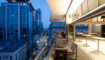 The image shows a modern balcony view of a city at dusk, with outdoor seating and tables, overlooking a street lined with tall buildings.