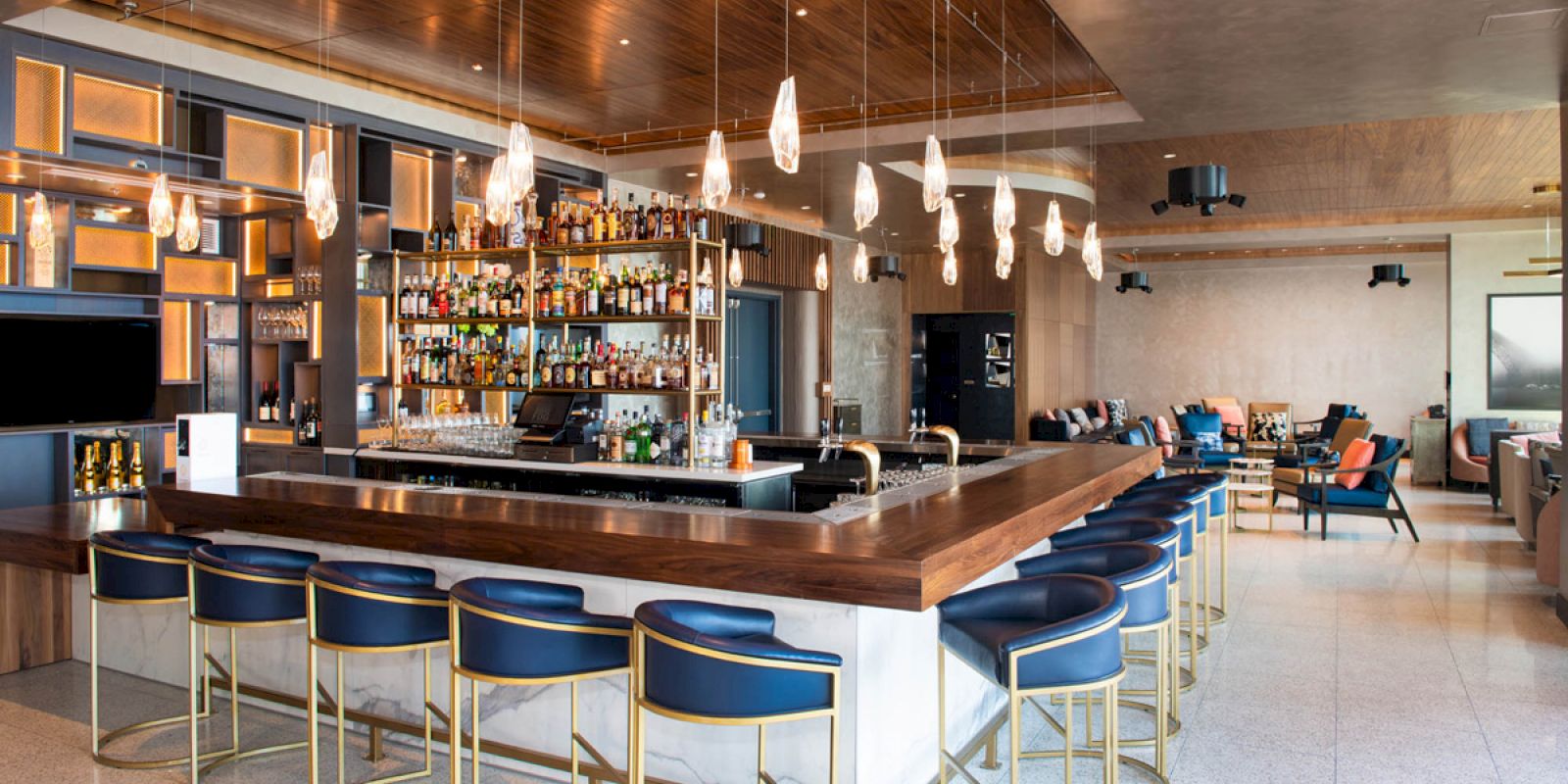 A modern bar with blue cushioned stools, a wooden countertop, hanging lights, and a well-stocked selection of bottles behind the bar.