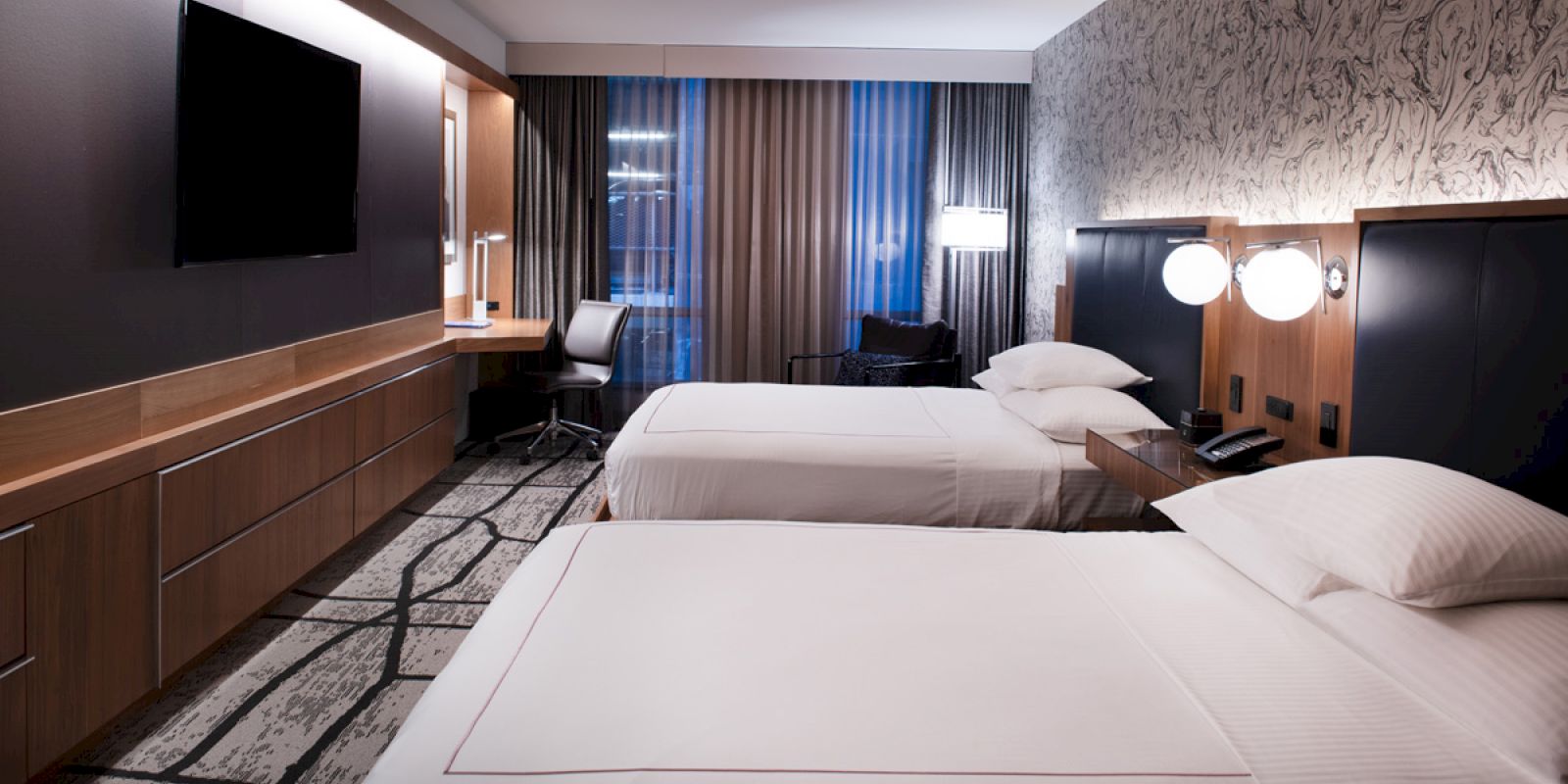 A modern hotel room with two neatly made beds, a wall-mounted TV, a desk with a chair, and contemporary decor.
