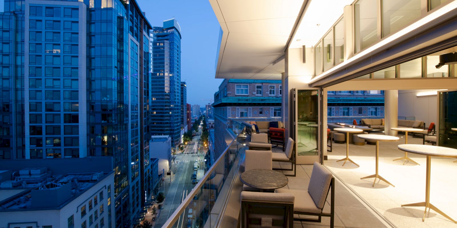 A modern city view from a balcony at dusk, featuring tall buildings, street lights, chairs, and small round tables arranged outside.