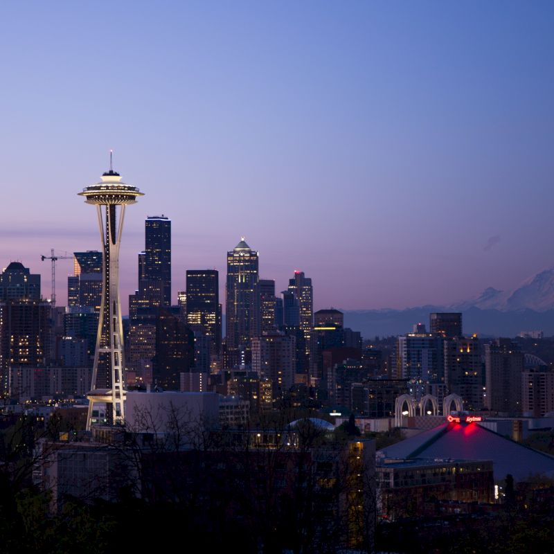 The image depicts the Seattle skyline at dusk, with the Space Needle prominently featured and Mount Rainier in the background.