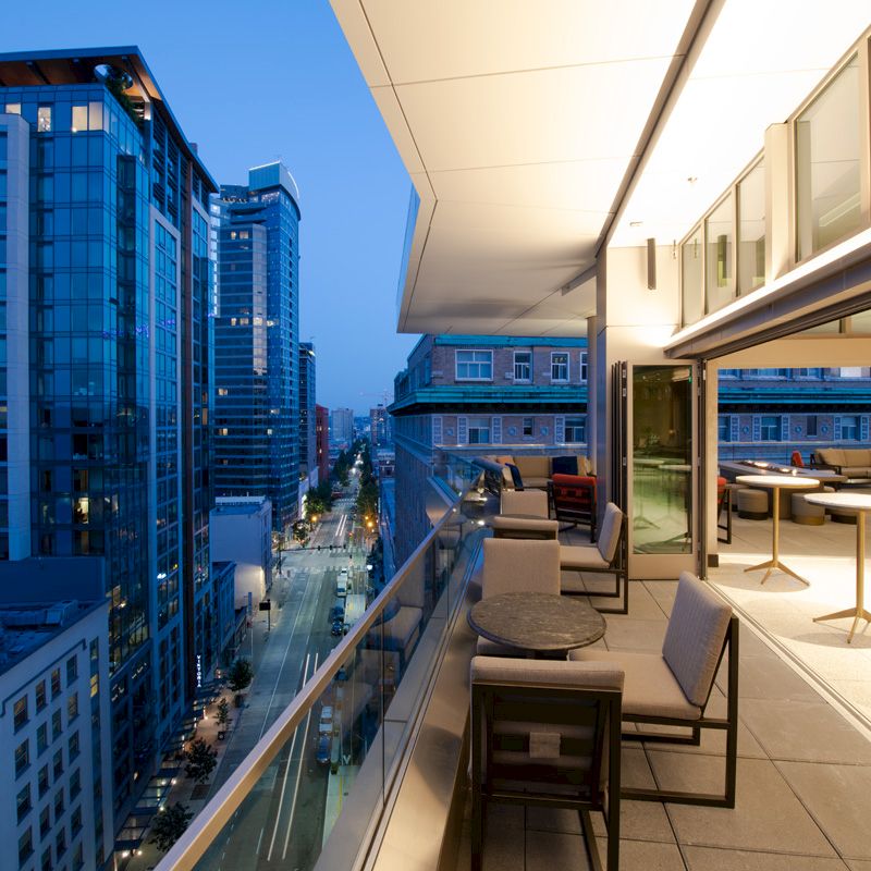 A modern balcony at twilight with seating areas overlooking a city street lined with tall buildings. The interior lights spill onto the balcony.