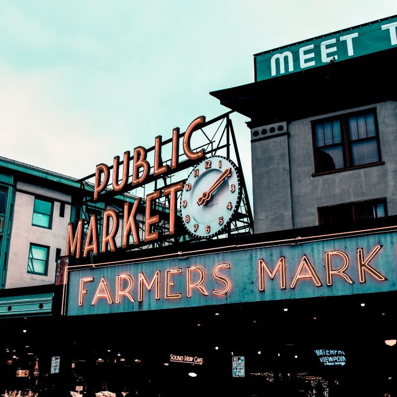 The image shows an iconic sign for a "Public Market" and "Farmers Market" with a clock, set against urban buildings, under a cloudy sky.