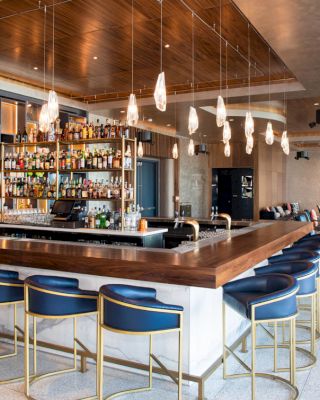 This image depicts a modern bar with a wooden counter, blue chairs, hanging lights, and a well-stocked shelf of bottles.