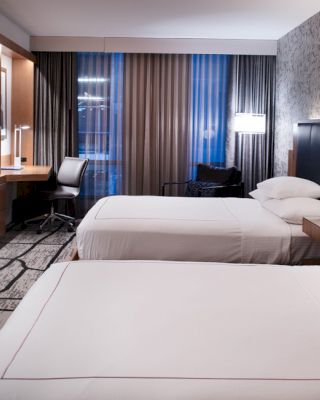 A modern hotel room with two beds, a wall-mounted TV, a work desk with a chair, and stylish decor including a patterned carpet and wallpaper.