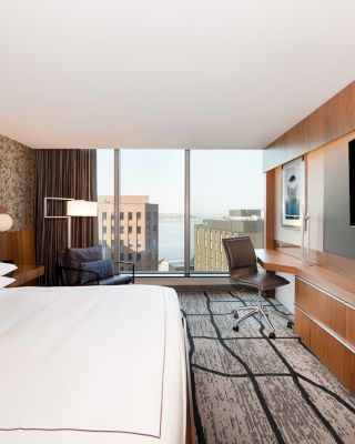A modern hotel room with a king-sized bed, desk, chair, large TV, and a view of city buildings through floor-to-ceiling windows.