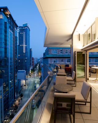 An outdoor patio of a modern building overlooking a city street lined with high-rise buildings at dusk, featuring tables and chairs.