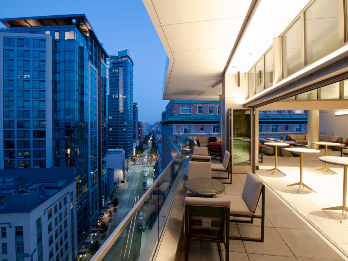 A modern cityscape seen from a high-rise balcony at dusk, featuring outdoor seating and city buildings with large glass windows.