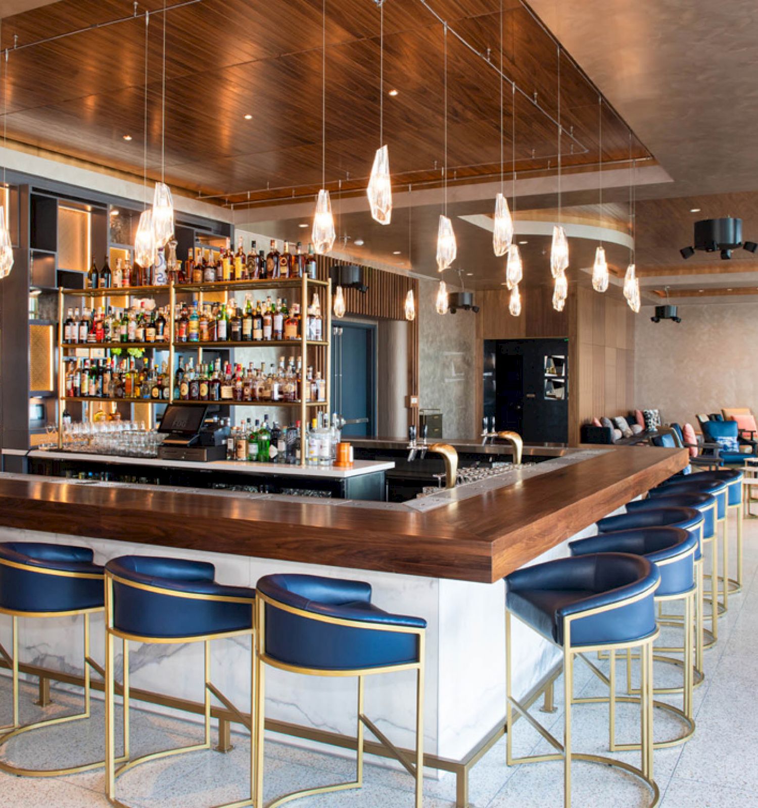 A modern bar with a central counter, blue bar stools, hanging light fixtures, and a well-stocked shelf with bottles. The seating area is visible.