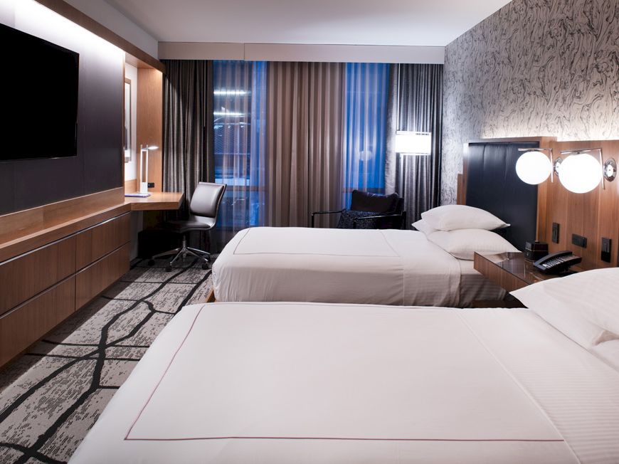 A modern hotel room features two neatly made beds, a flat-screen TV, a desk with a chair, and wall decor, bathed in soft lighting.