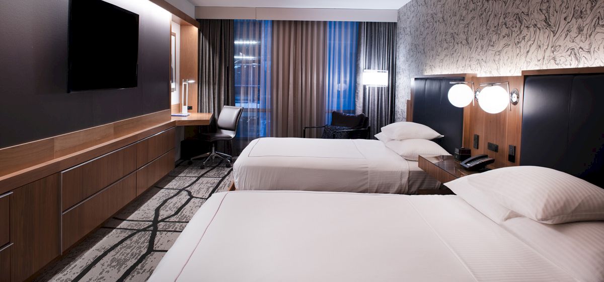 A modern hotel room features two neatly made beds, a flat-screen TV, a desk with a chair, and wall decor, bathed in soft lighting.