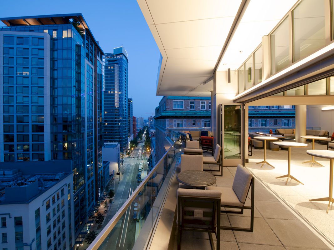 A modern cityscape viewed from a balcony at dusk, featuring high-rise buildings and a furnished outdoor seating area with tables and chairs.