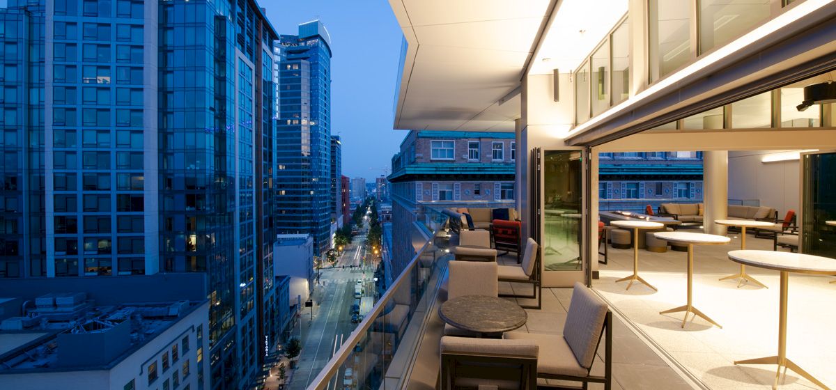 A modern cityscape viewed from a balcony at dusk, featuring high-rise buildings and a furnished outdoor seating area with tables and chairs.