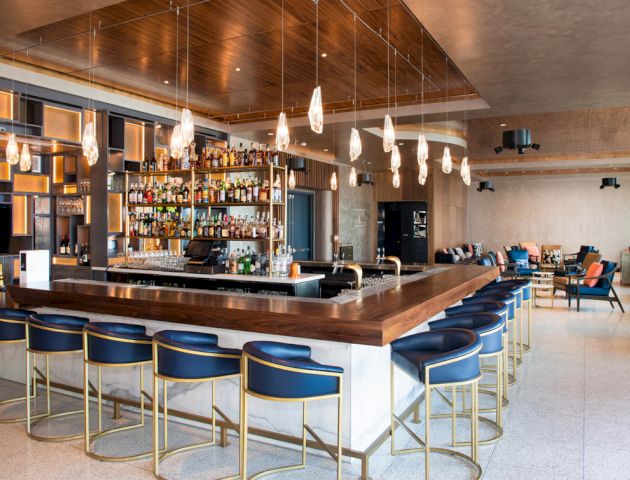 The image shows a modern bar with a long counter, blue stools, hanging lights, and a well-stocked liquor shelf in a spacious, stylish interior.