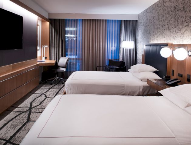 A modern hotel room with two double beds, a wall-mounted TV, a desk with a chair, and large windows with curtains and a view outside.