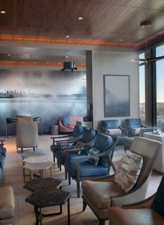 A modern lounge with various chairs, small tables, and large windows offering a view of a city skyline and water beyond.