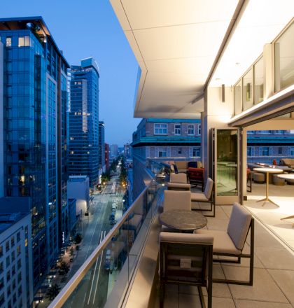 This image shows a modern rooftop terrace with tables and chairs overlooking a city street lined with tall buildings at dusk.