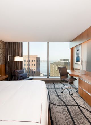 A modern hotel room with a king-sized bed, desk, TV, and large window offering a city view. Neutral tones and contemporary furniture fill the space.