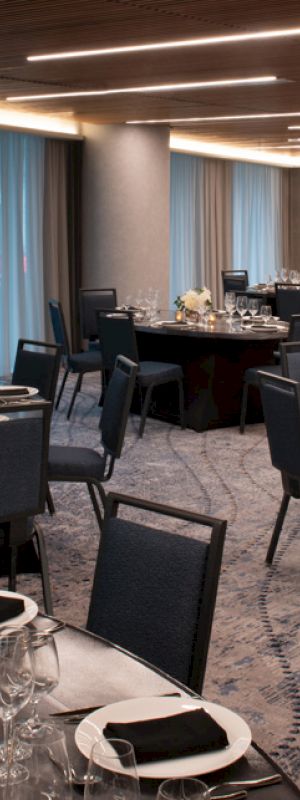 This image shows a modern, elegantly set dining hall with round tables adorned with white flowers and black napkins on silverware, ready for an event.