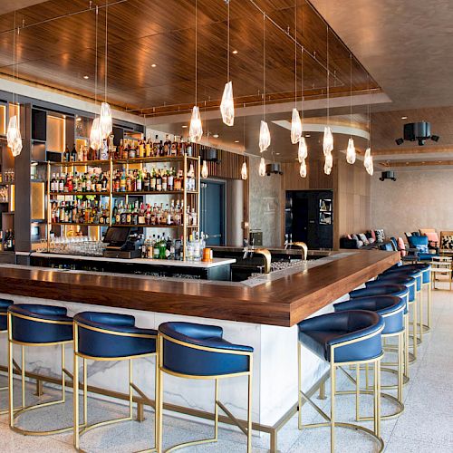 A modern bar with blue stools, a variety of drinks displayed on shelves, hanging lights, and a comfortable seating area in the background.