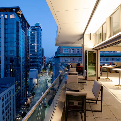 An open high-rise terrace with seating, overlooking a cityscape with illuminated buildings at dusk. The terrace has tables and chairs arranged neatly.