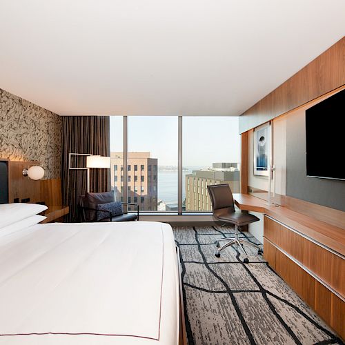 A modern hotel room with a large bed, a desk with a chair, a wide window showing buildings, and a wall-mounted TV.