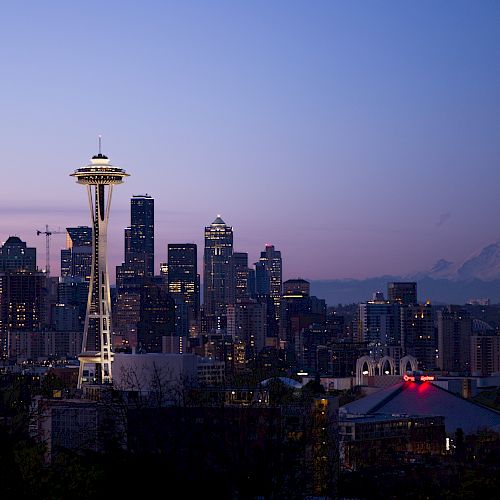 A cityscape with a prominent tower at dusk, with a mountain in the background, and an arena with a red top in the lower right corner of the image.