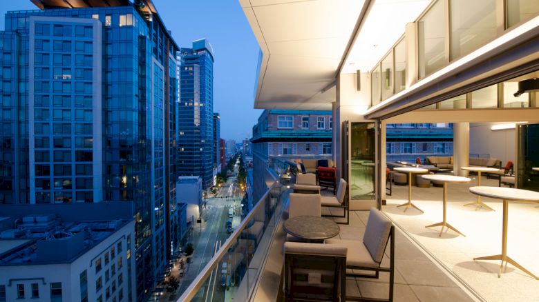 A modern cityscape featuring tall buildings and a well-lit balcony with tables and chairs at night, overlooking a busy street.