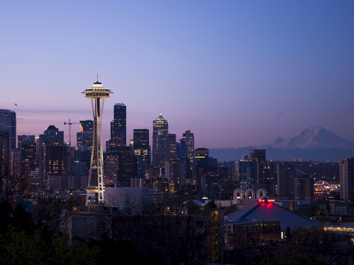 A city skyline at dusk featuring a prominent tower (Space Needle), with tall buildings illuminated and a distant mountain (Mount Rainier).
