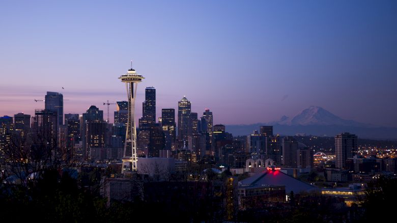 A city skyline at dusk featuring a prominent tower (Space Needle), with tall buildings illuminated and a distant mountain (Mount Rainier).