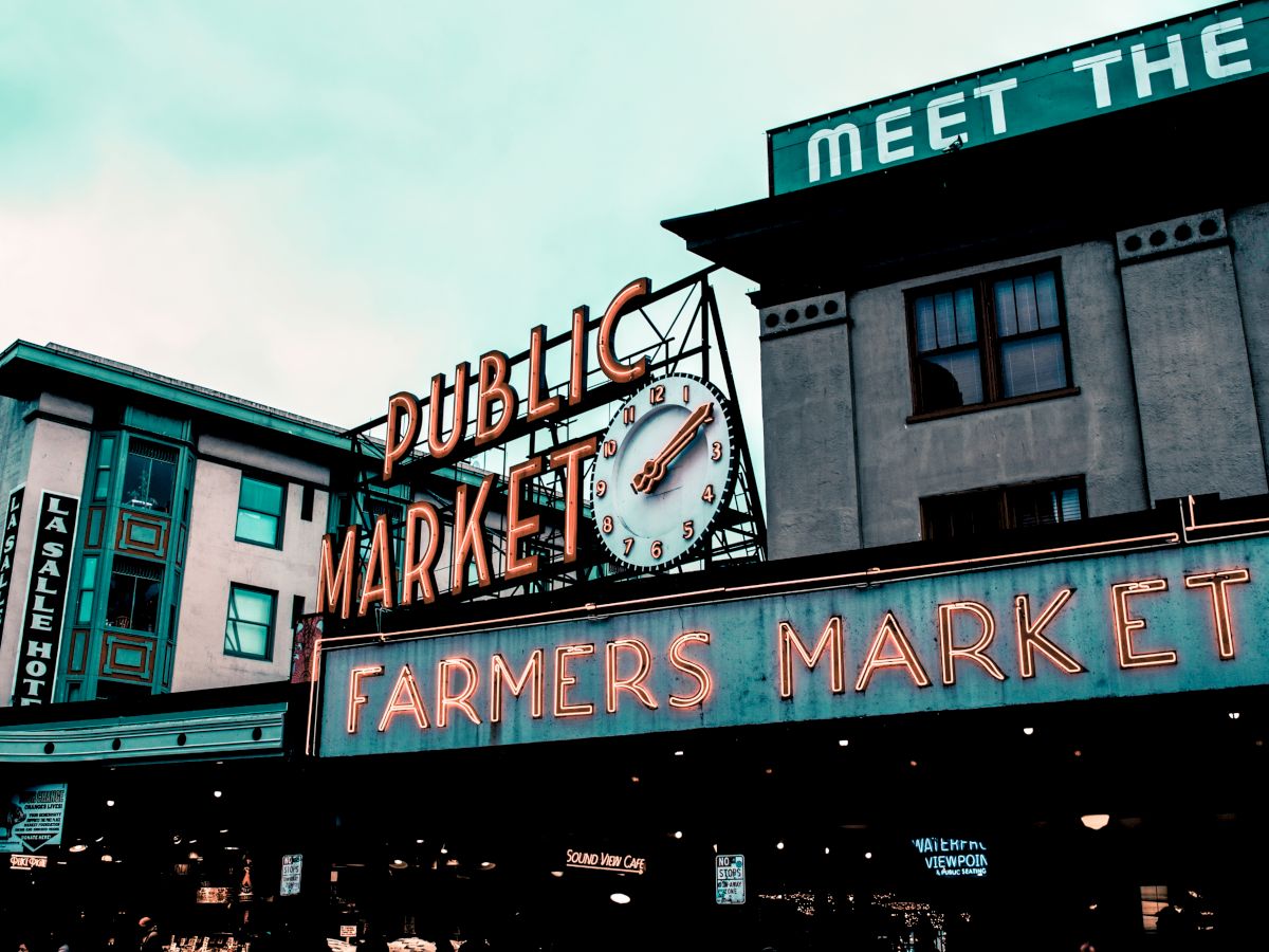 The image shows a neon sign for a "Public Market" with a clock, and below it, another sign for a "Farmers Market" on a building.