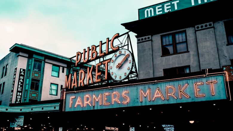 The image shows a neon sign for a "Public Market" with a clock, and below it, another sign for a "Farmers Market" on a building.